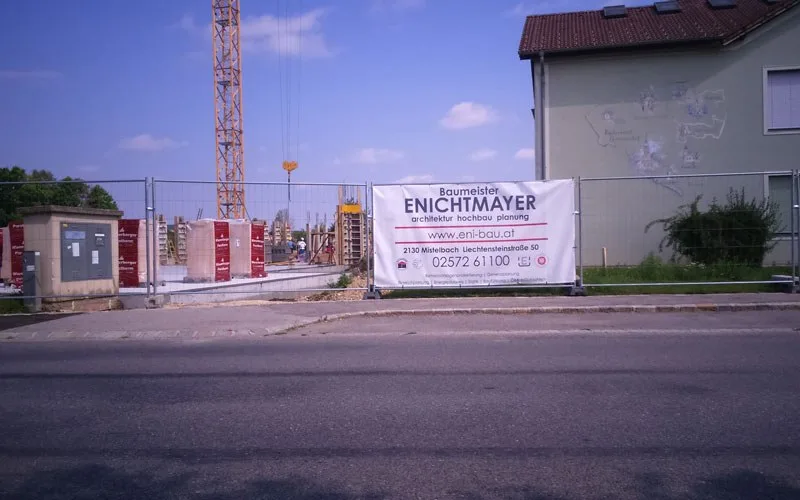 Baustelle mit Kran, davor ein Banner von Baumeister Enichtmayer am Bauzaun